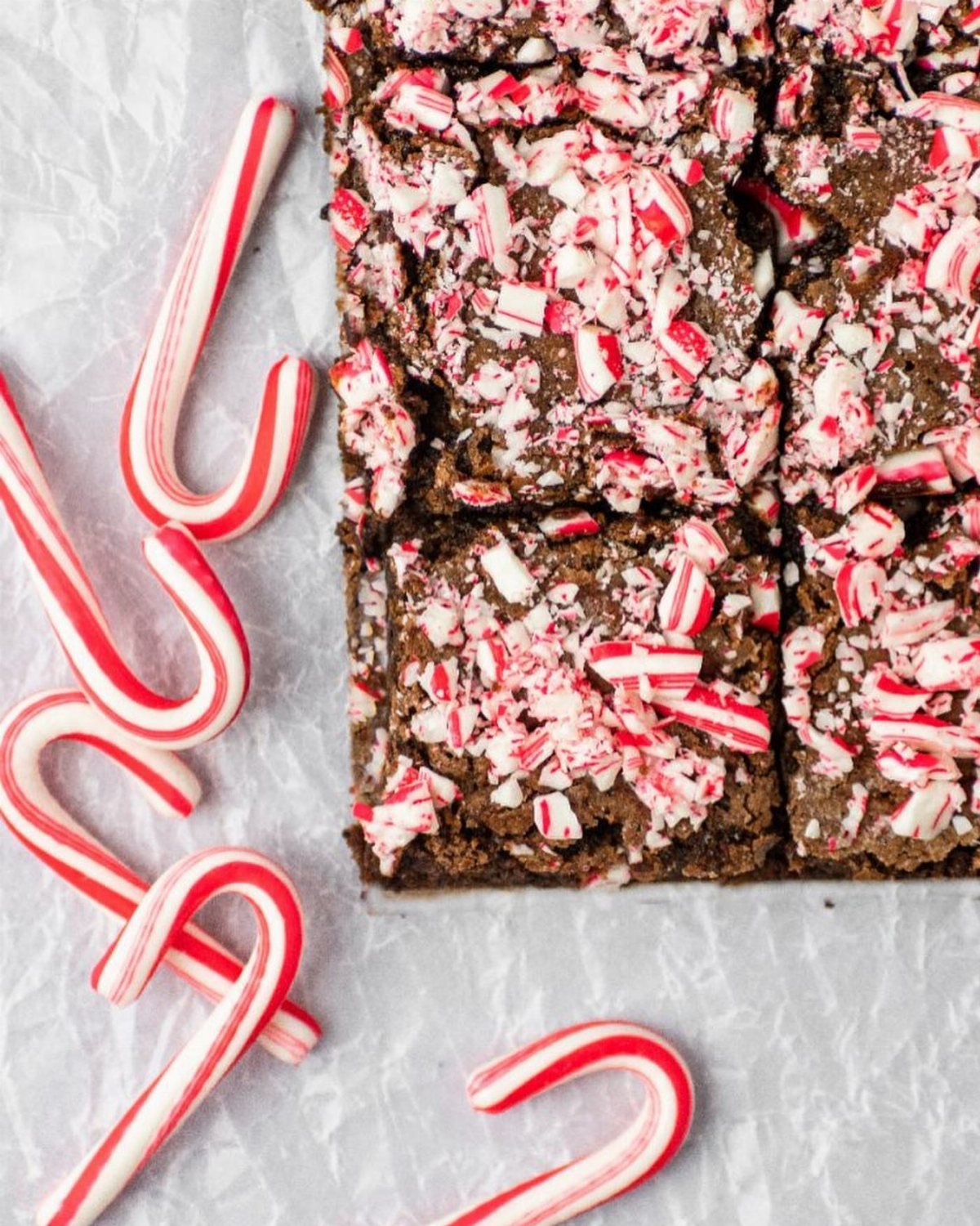 Chocolate brownies topped with crushed candy cane pieces on white parchment paper with whole candy canes.