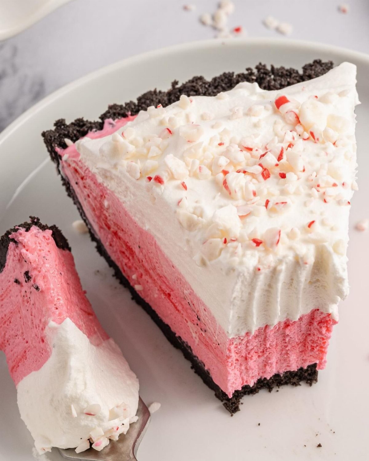 A slice of pink peppermint pie with whipped cream and crushed candy cane pieces on a plate.