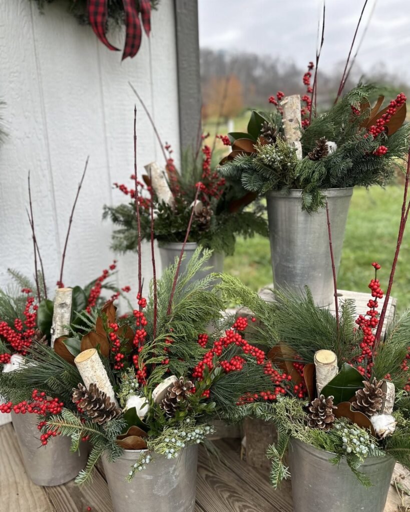 Galvanized metal buckets filled with evergreen branches, red berries, and pinecones arranged on a wooden surface.