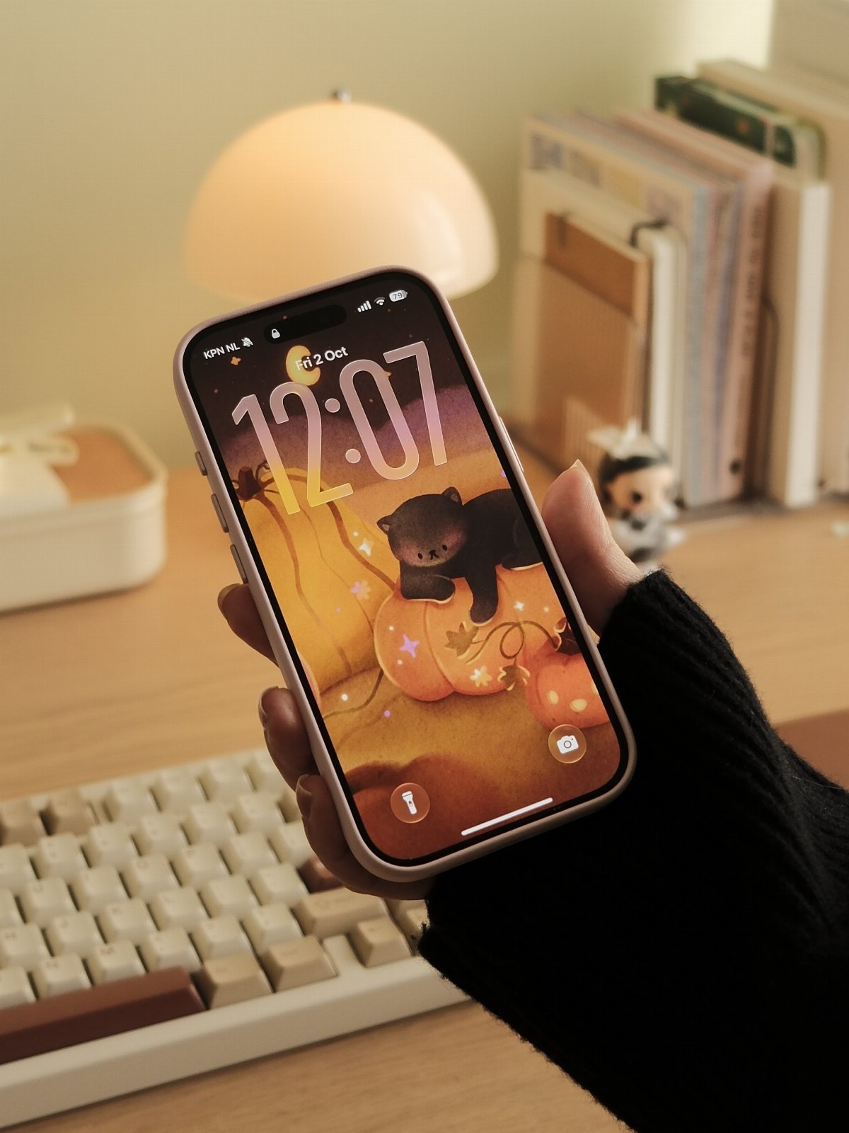 A hand holding a smartphone displaying a cute Halloween wallpaper with a black cat and pumpkins.