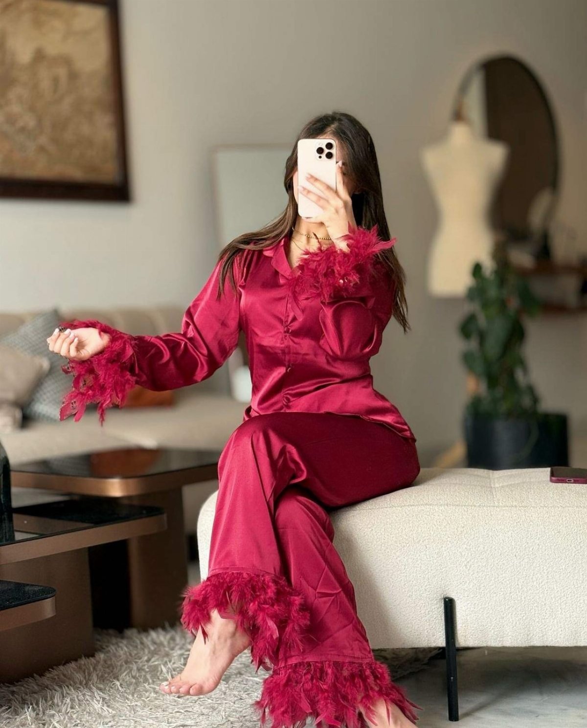 Woman in red satin pajamas with feather trim sitting on a white ottoman.