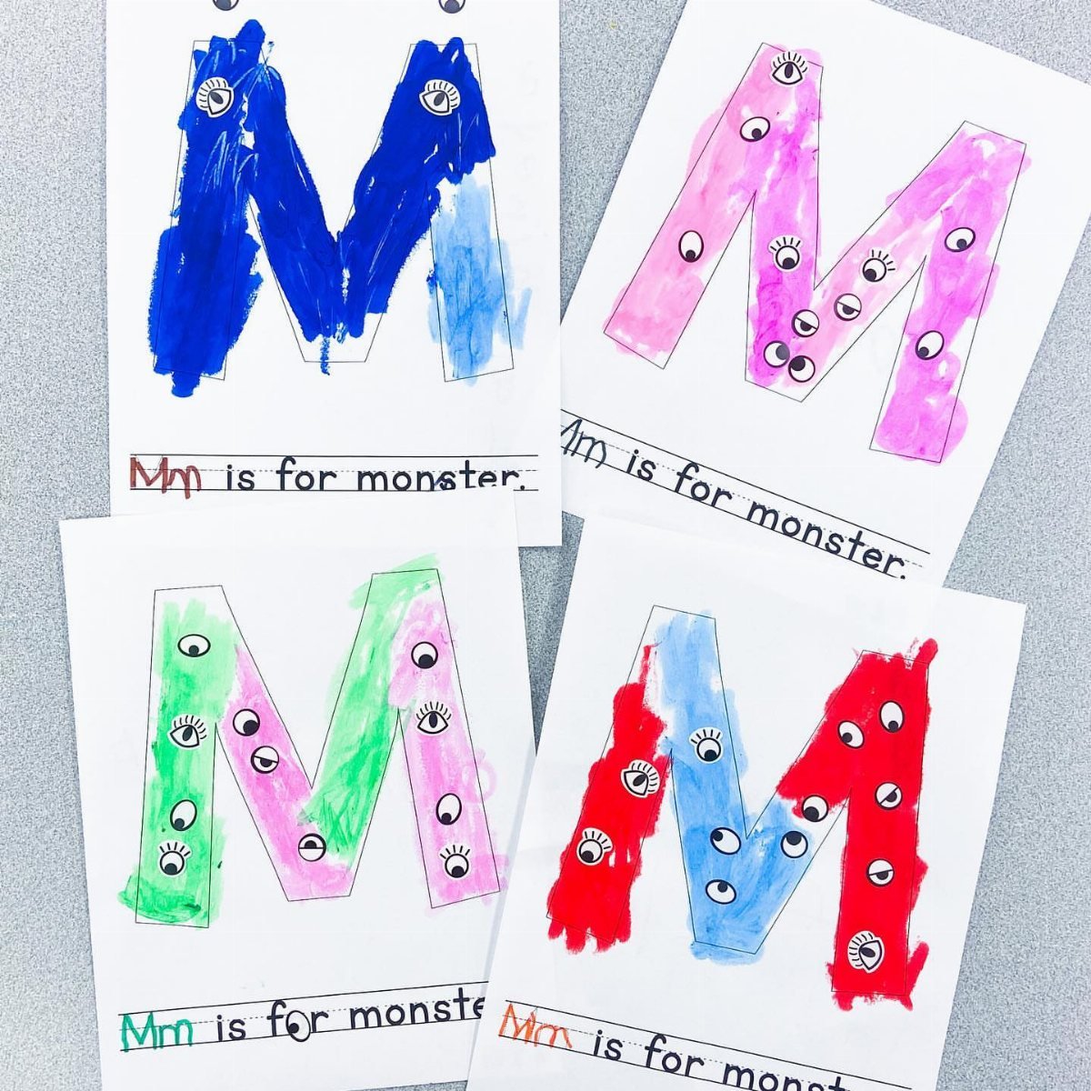 Four colorful letter M monster art pages with googly eyes on a grey surface.