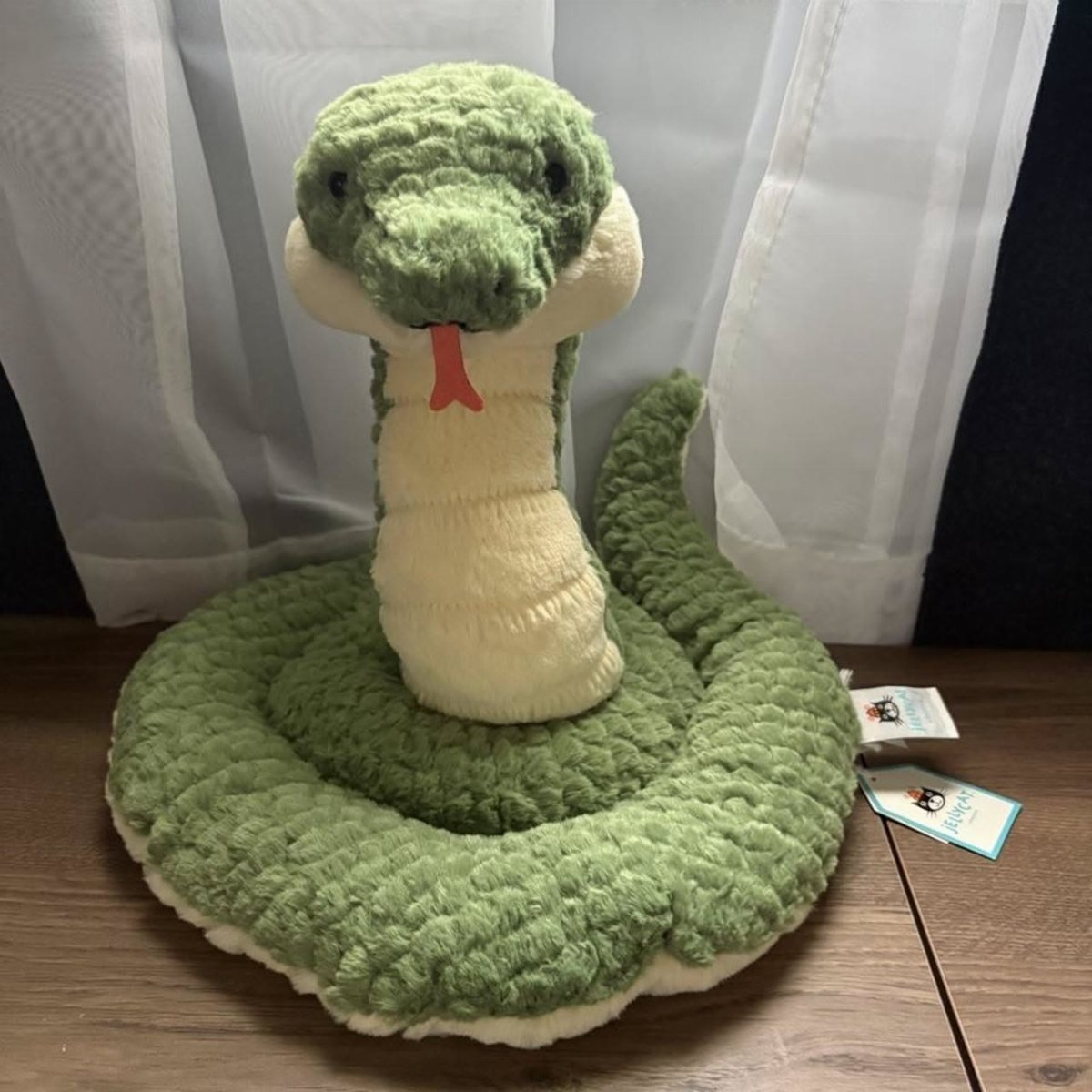 A green and cream stuffed snake toy with a red tongue sitting on a wooden surface.